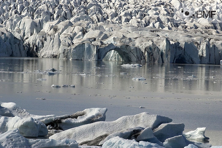 A striking image of a beautiful glacier. The ice and its varied shapes frame the upper and lower part of the photograph. From left to right, a strip of still sea water merges with its white and silver walls.