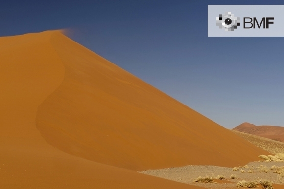 Una impactant duna de sorra ataronjada i vermellosa protagonitza aquesta imatge. El fotògraf aconsegueix captar l'instant en que una ràfega d'aire s'enduu la sorra del seu punt més alt i es mescla amb el blau intens del cel.
